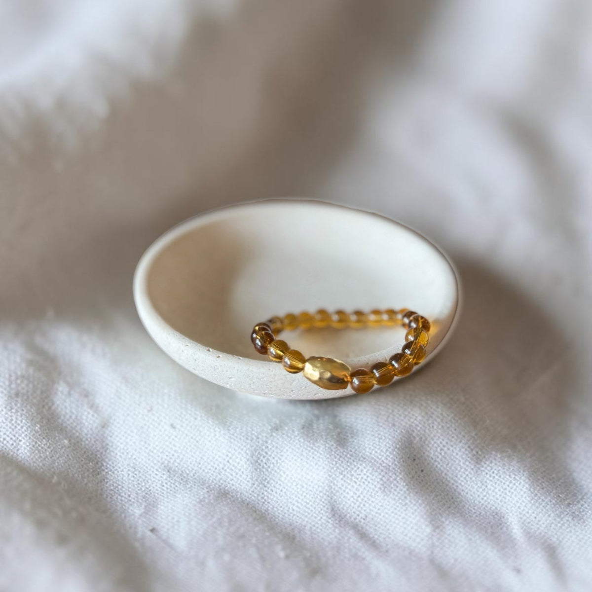 A beaded stretch bracelet with amber-brown translucent beads and a gold-toned clasp, resting inside a small white ceramic dish on a soft white fabric background.