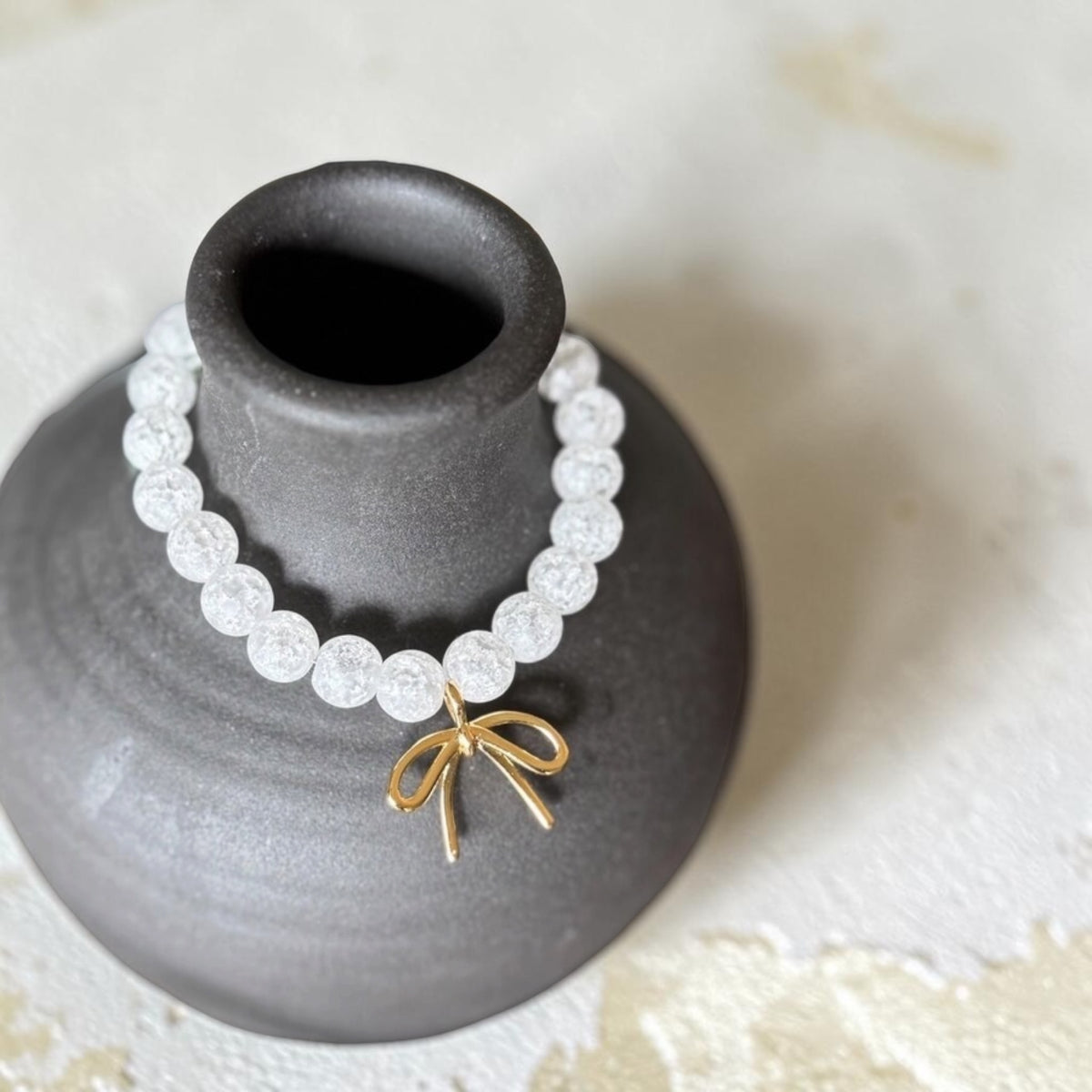 A white beaded bracelet with a small gold bow charm, displayed on a dark gray ceramic vase against a light textured background.