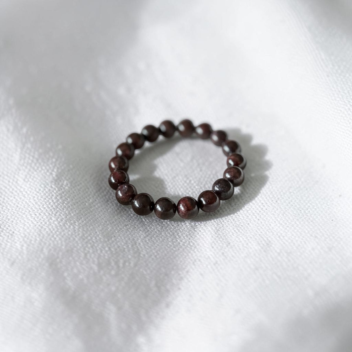 A beaded stretch bracelet made of small, polished dark brown wooden beads, resting on a textured white fabric background.