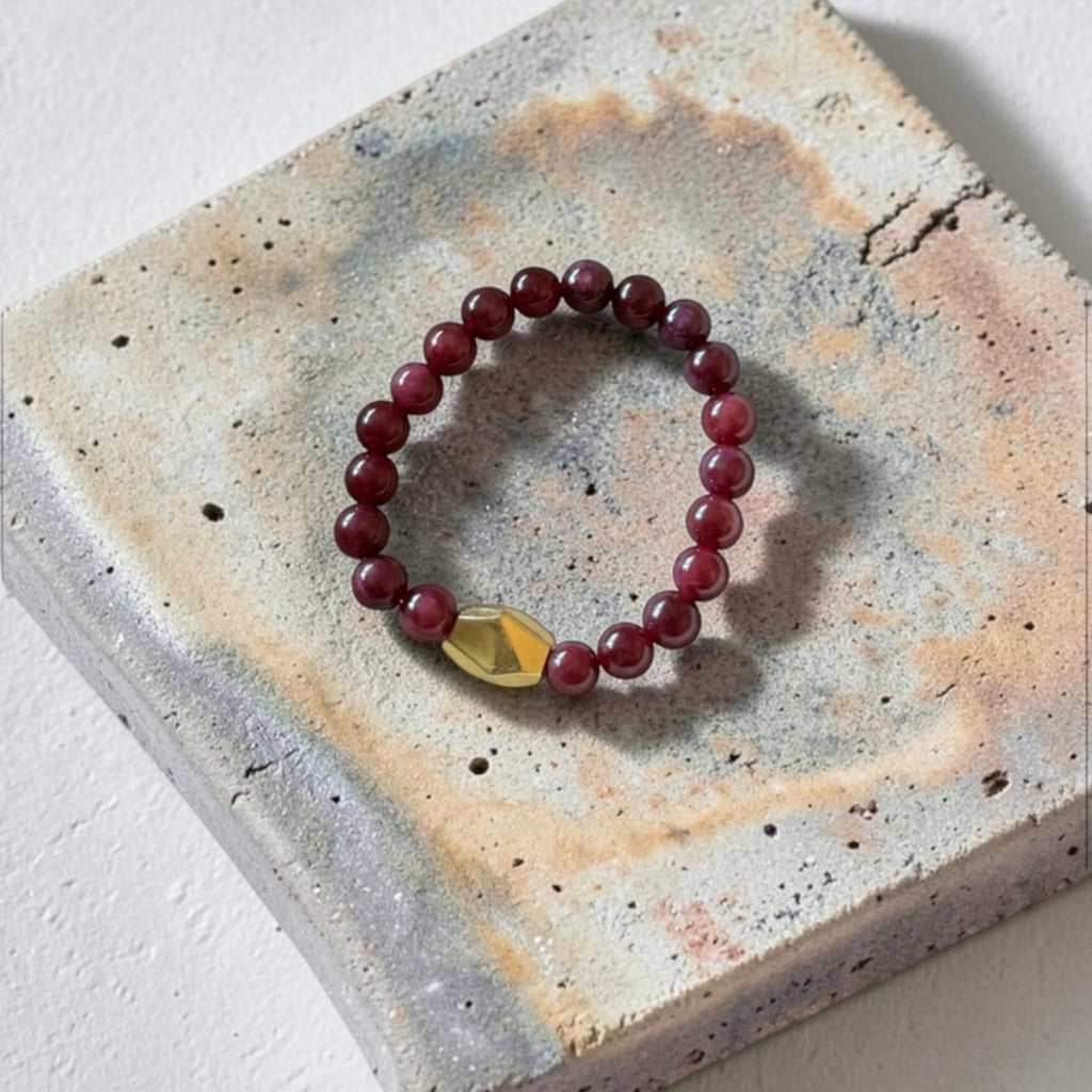 A beaded stretch bracelet with dark red round beads and a faceted gold accent bead, displayed on a textured concrete tile against a white background.