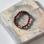 A beaded stretch bracelet with dark red round beads and a faceted gold accent bead, displayed on a textured concrete tile against a white background.