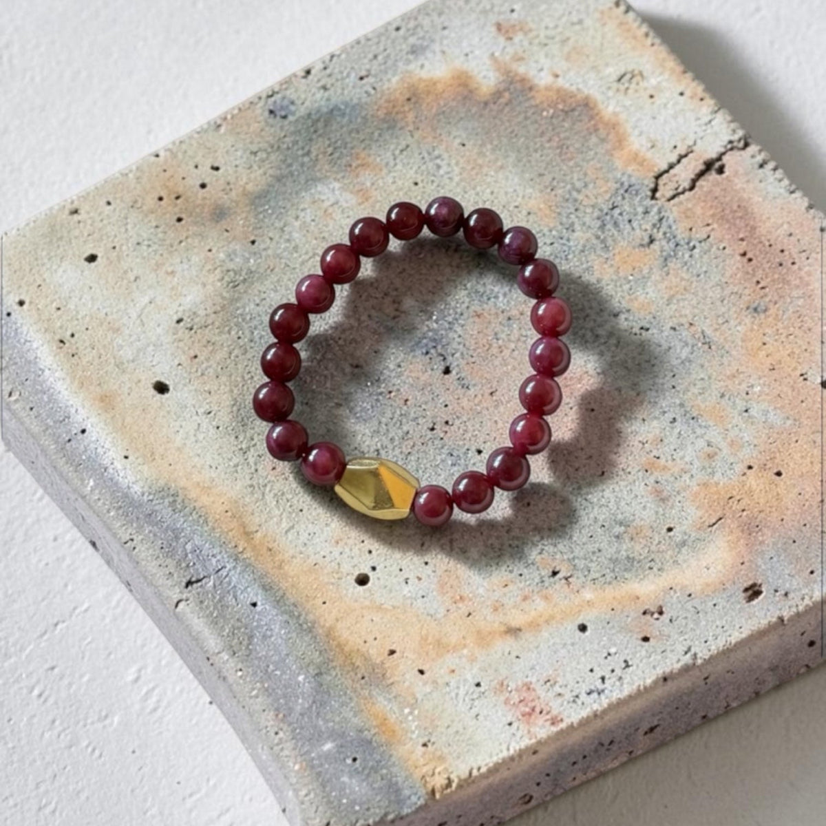 A beaded stretch bracelet with dark red round beads and a faceted gold accent bead, displayed on a textured concrete tile against a white background.