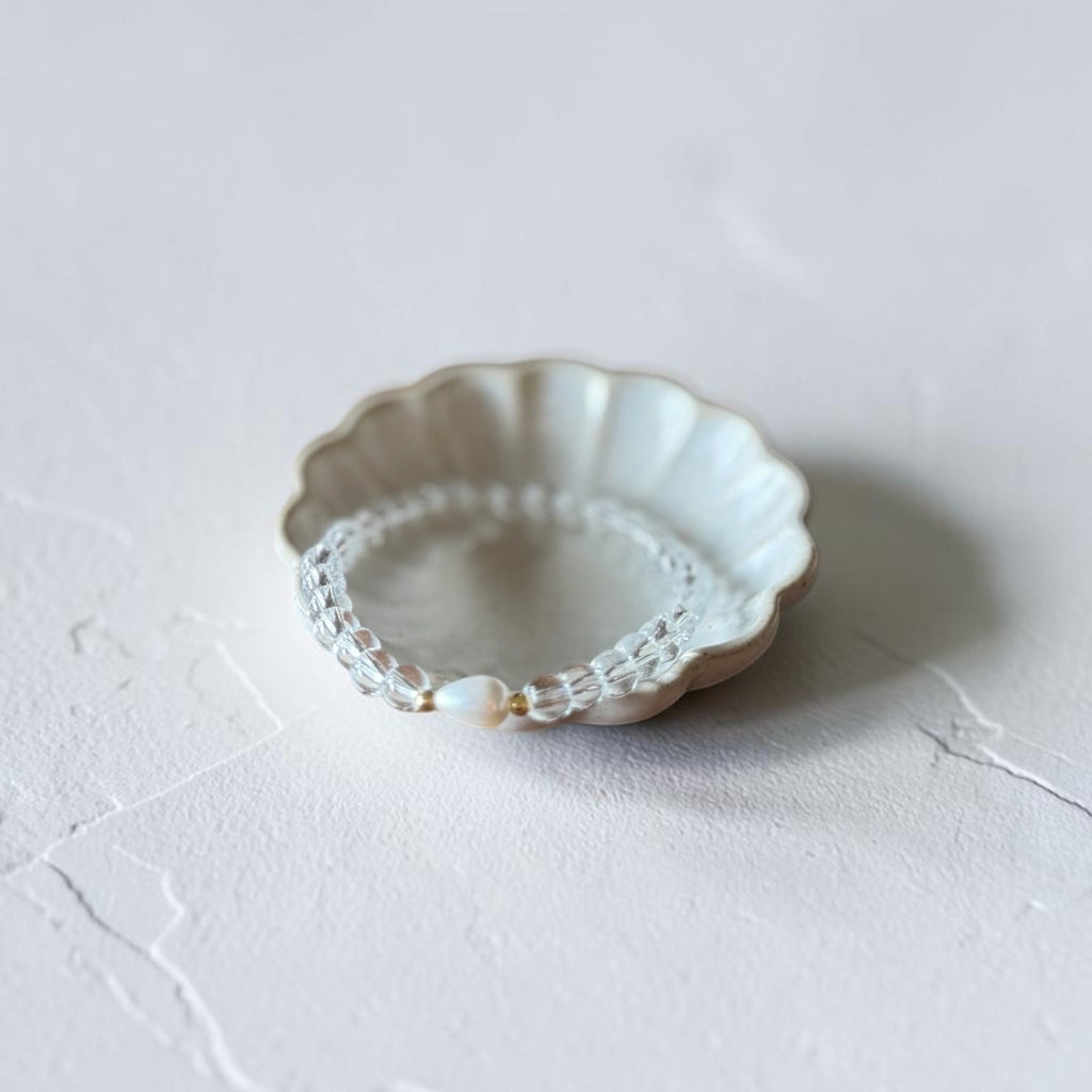 A delicate clear beaded bracelet with a central pearl and gold-toned accents, displayed inside a scalloped ceramic dish on a textured light gray surface.
