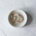 Three delicate beaded stretch bracelets in soft pink, white, and gold tones, displayed inside a small speckled ceramic dish on a textured light gray surface.
