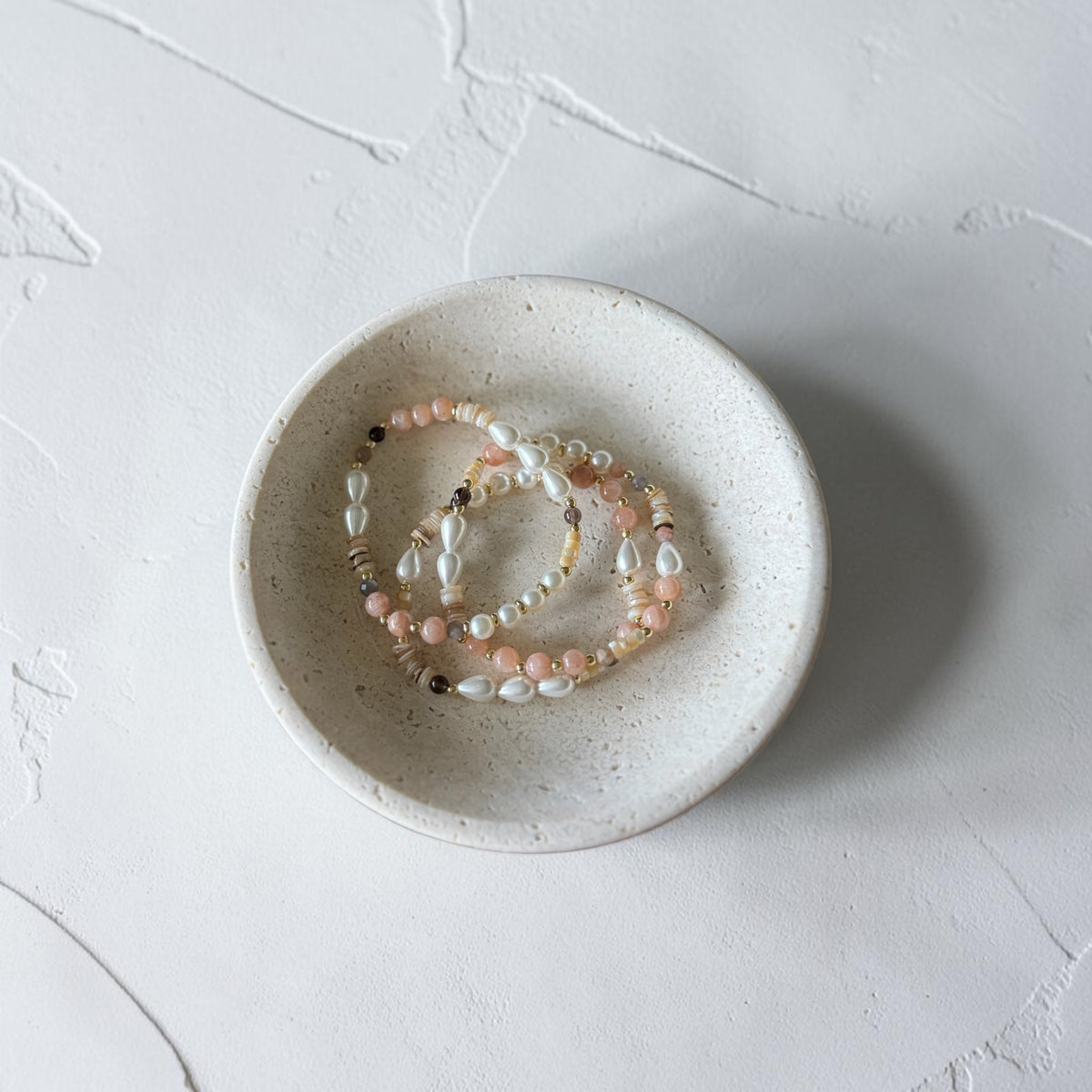 Three delicate beaded stretch bracelets in soft pink, white, and gold tones, displayed inside a small speckled ceramic dish on a textured light gray surface.
