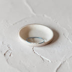 A thin dark teal seed bead wire bracelet, displayed inside a small, round, handmade ceramic dish on a textured light gray surface.