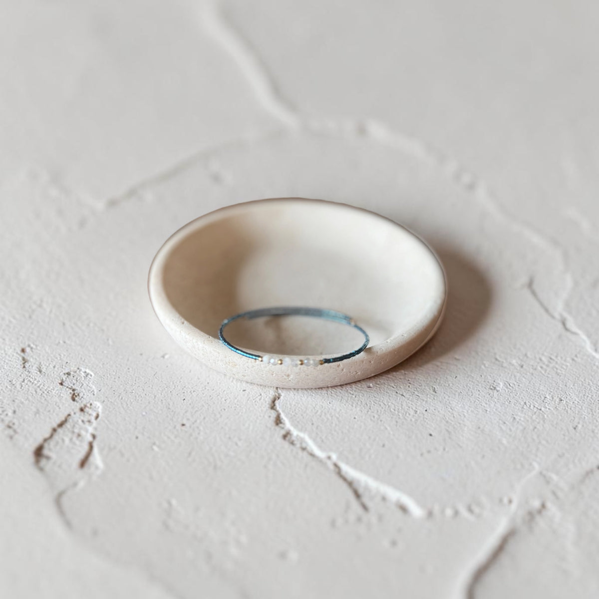 A thin dark teal seed bead wire bracelet, displayed inside a small, round, handmade ceramic dish on a textured light gray surface.