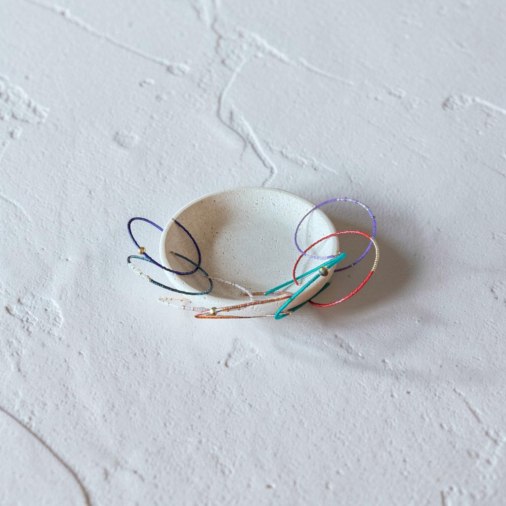 A small white ceramic dish or shallow bowl on a textured white surface, with several thin multicolored wire loops or rings draped around and inside it.