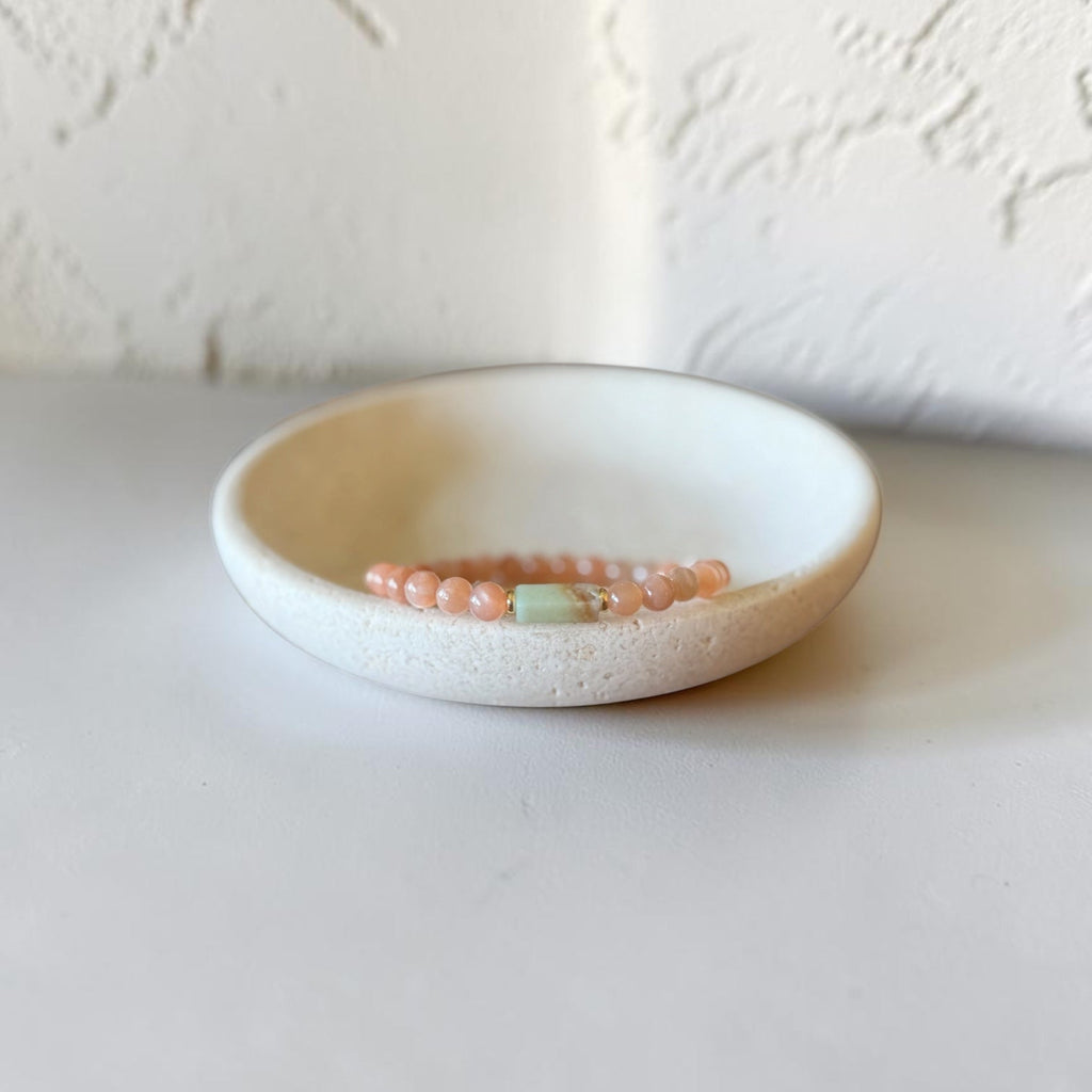 A beaded bracelet with peach-pink round beads and a central mint-green spacer bead, displayed on a small round ceramic dish against a textured off-white background.