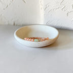 A beaded bracelet with peach-pink round beads and a central mint-green spacer bead, displayed on a small round ceramic dish against a textured off-white background.