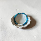 A beaded stretch bracelet in shades of blue with a small white pearl accent, displayed inside a small white ceramic dish on a textured off-white surface.