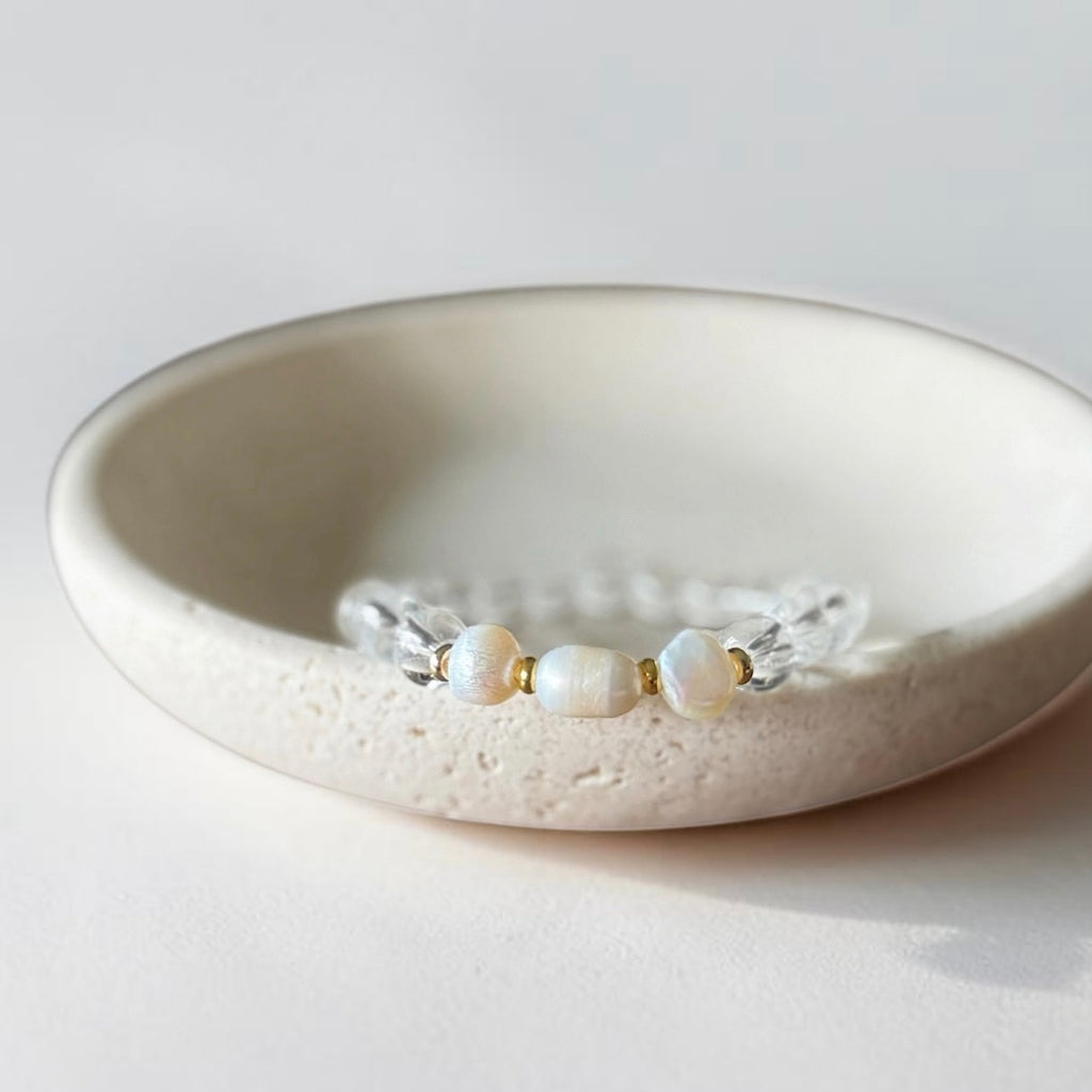A delicate beaded bracelet with three white pearl-like beads and clear faceted beads, displayed inside a textured cream ceramic dish on a light neutral background.