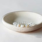 A delicate beaded bracelet with three white pearl-like beads and clear faceted beads, displayed inside a textured cream ceramic dish on a light neutral background.