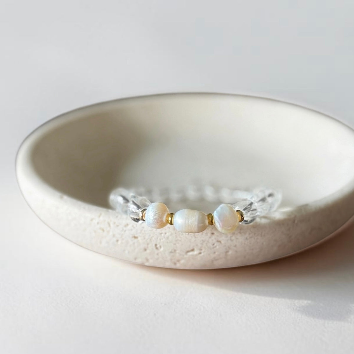 A delicate beaded bracelet with three white pearl-like beads and clear faceted beads, displayed inside a textured cream ceramic dish on a light neutral background.