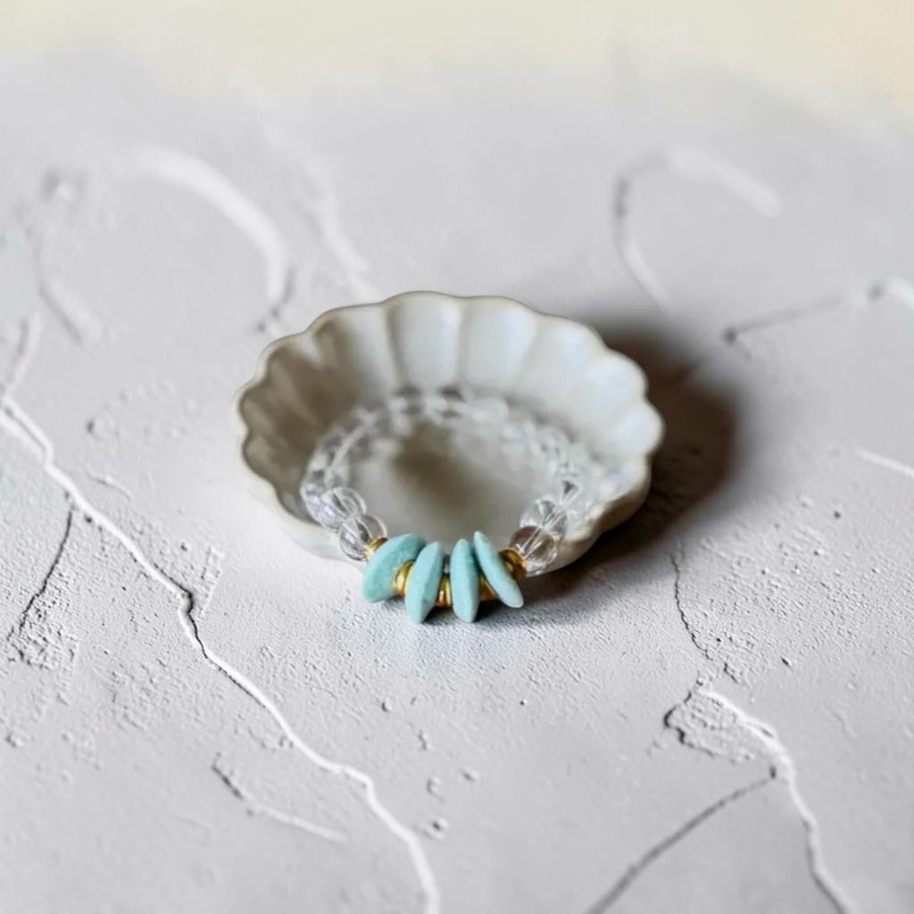 A beaded ring with light blue teardrop beads and clear faceted beads, displayed on a small white scalloped dish over a textured light gray surface.