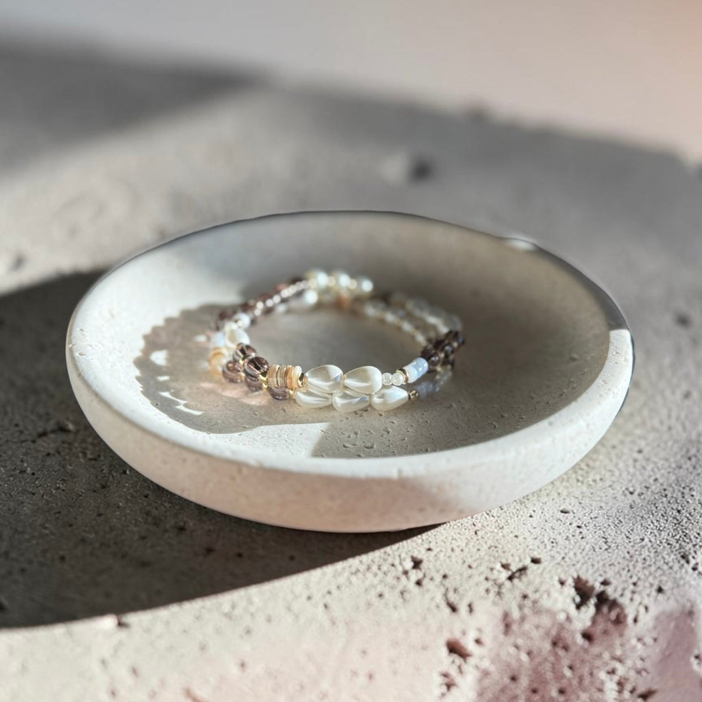 A delicate beaded bracelet with white pearls and small gold-toned beads, displayed on a small round ceramic dish over a textured concrete surface.