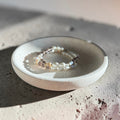 A delicate beaded bracelet with white pearls and small gold-toned beads, displayed on a small round ceramic dish over a textured concrete surface.
