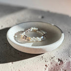 A delicate beaded bracelet with white pearls and small gold-toned beads, displayed on a small round ceramic dish over a textured concrete surface.