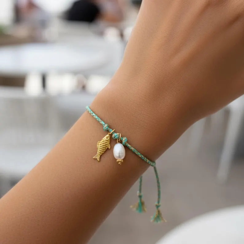 A close-up of a person's wrist wearing a delicate mint green cord bracelet with gold charms, including a fish and a pearl bead.