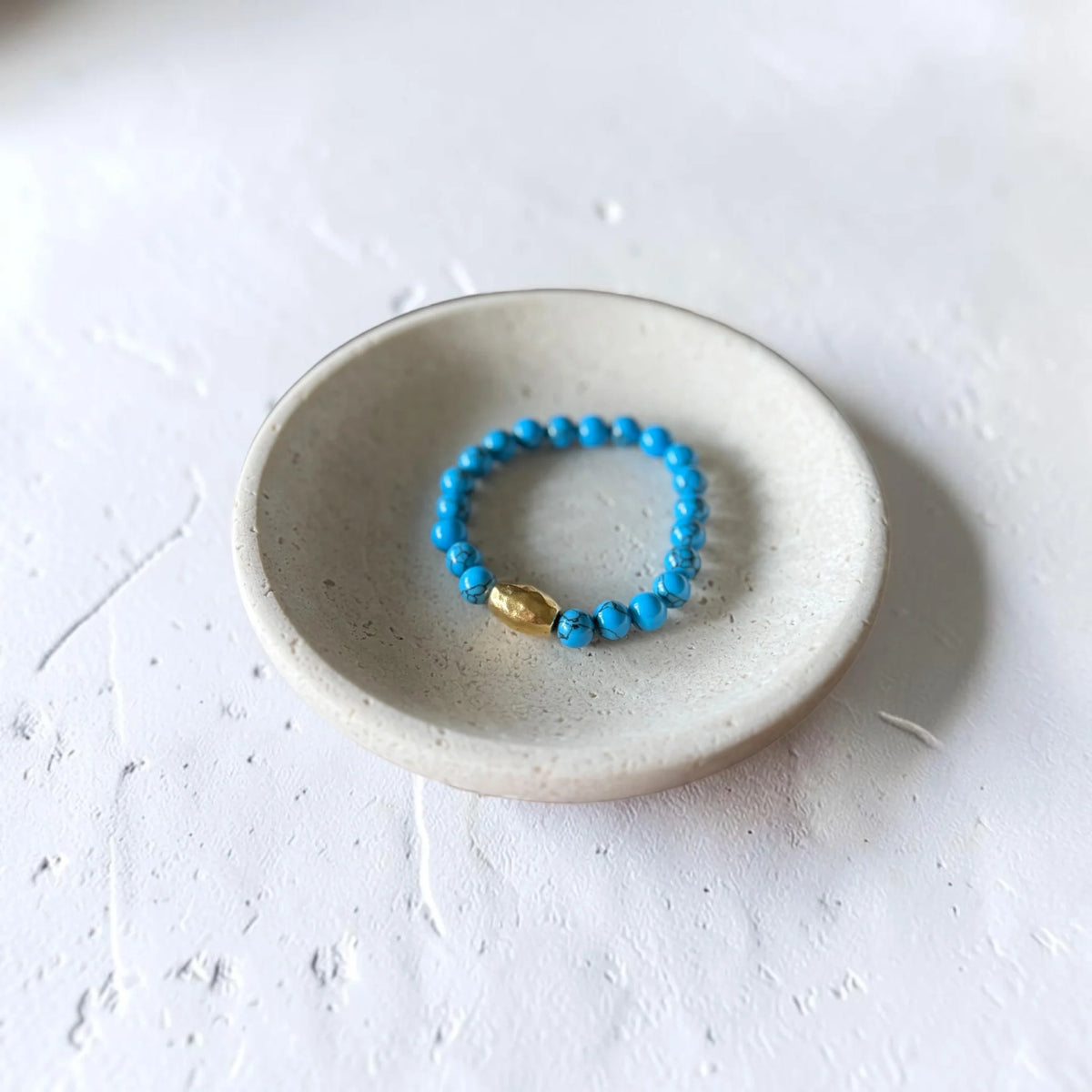 A beaded stretch ring with bright blue beads and a small gold accent bead, displayed inside a small speckled ceramic dish on a textured white surface.