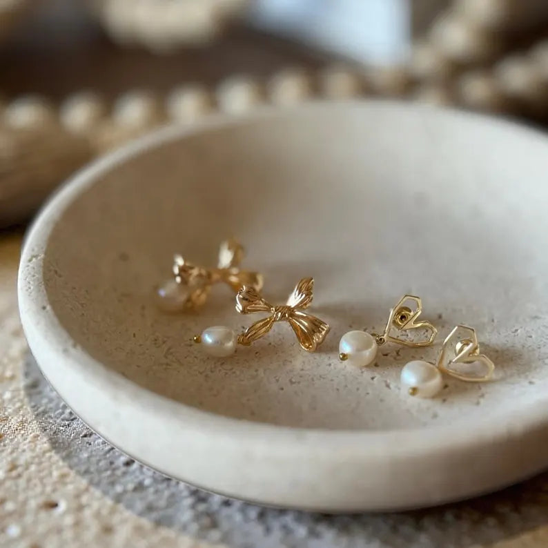 A pair of gold bow stud earrings with small white pearl drops and a pair of gold heart-shaped wire earrings with pearl studs, displayed on a round beige stone dish.