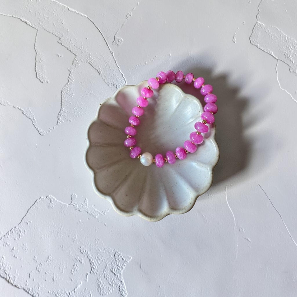 A pink beaded stretch bracelet with a single white pearl accent, displayed on a scalloped white ceramic dish against a textured white background.