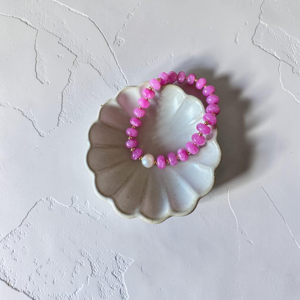 A pink beaded stretch bracelet with a single white pearl accent, displayed on a scalloped white ceramic dish against a textured white background.