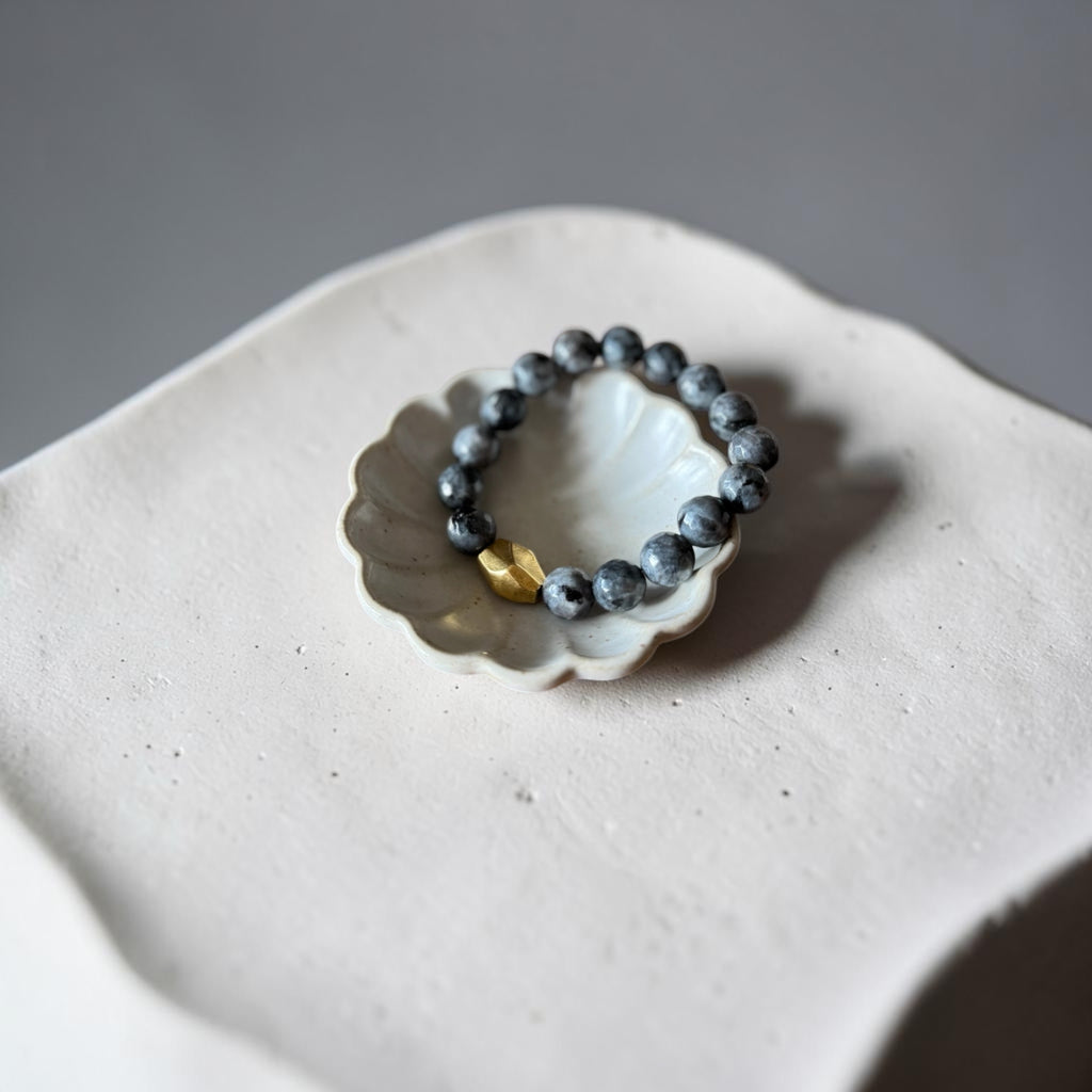 A beaded bracelet with dark gray stone beads and a small gold accent bead, displayed on a white ceramic shell-shaped dish against a neutral gray background.
