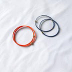 Two thin wrap-style bracelets on a white fabric background: one bright orange-red and one dark blue, each with small metallic gold bead accents.