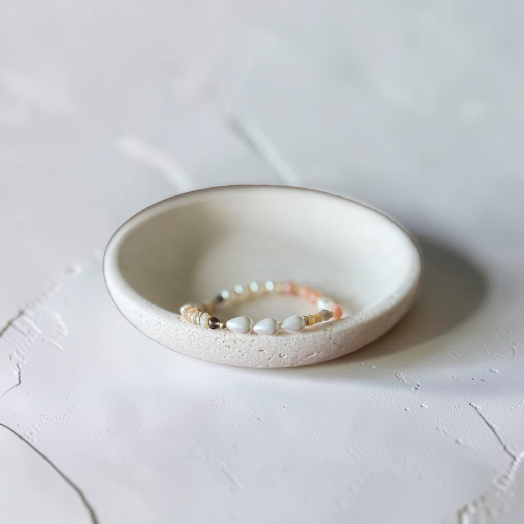 A delicate beaded bracelet with white, pink, and gold beads resting inside a small, textured off-white ceramic dish on a light gray surface.
