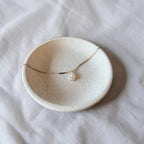 A delicate gold chain necklace with a single white pearl pendant, displayed on a small speckled ceramic dish over a white fabric background.