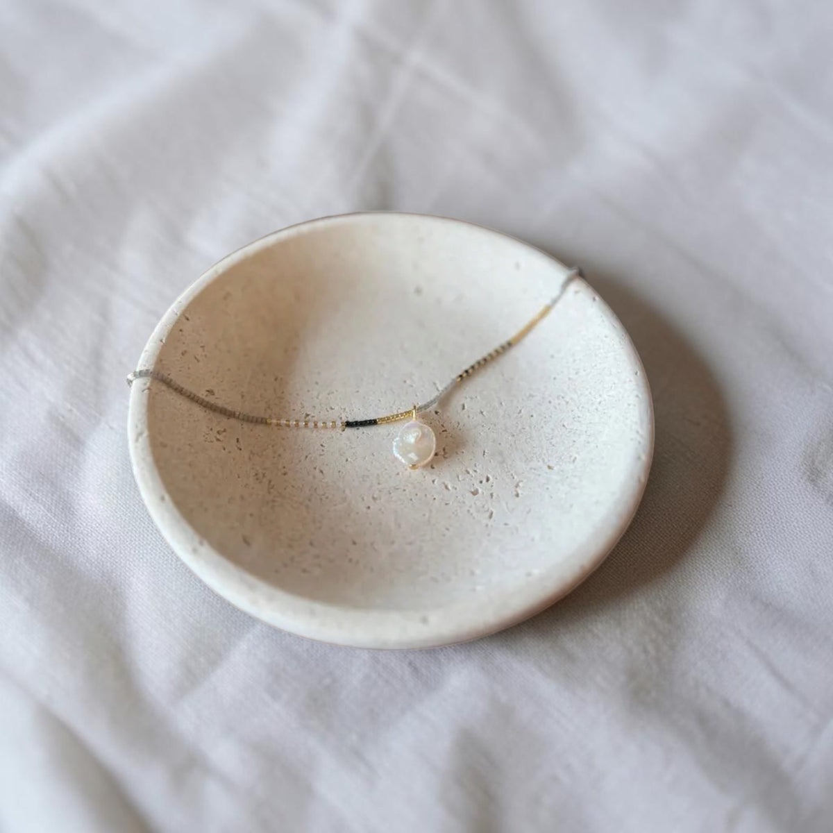 A delicate gold chain necklace with a single white pearl pendant, displayed on a small speckled ceramic dish over a white fabric background.