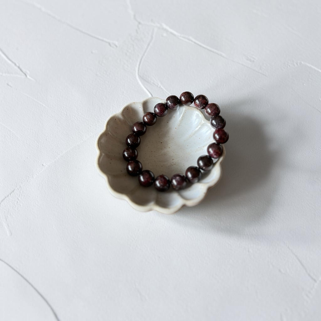 A beaded bracelet made of dark reddish-brown polished beads, displayed inside a small white ceramic dish on a textured white surface.