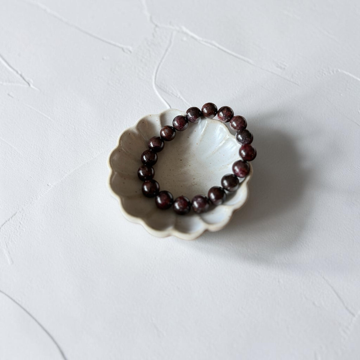 A beaded bracelet made of dark reddish-brown polished beads, displayed inside a small white ceramic dish on a textured white surface.