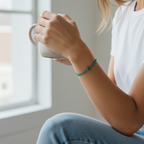 A woman in a white t-shirt and blue jeans sits by a window, holding a white ceramic mug. She is wearing a thin teal cord bracelet with small dark beads.