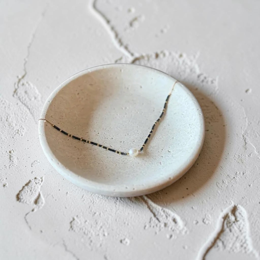 A delicate dark blue and white beaded necklace with a small white bead at the center, displayed on a shallow, round, speckled off-white ceramic dish against a textured, light beige stone background.