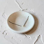 A delicate dark blue and white beaded necklace with a small white bead at the center, displayed on a shallow, round, speckled off-white ceramic dish against a textured, light beige stone background.