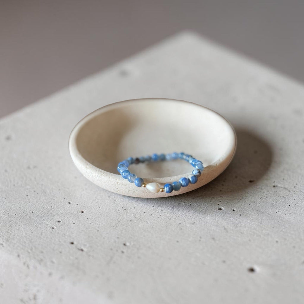 A beaded stretch bracelet with light blue beads and a small gold accent, displayed inside a small, round, off-white ceramic dish on a textured concrete surface.