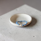 A beaded stretch bracelet with light blue beads and a small gold accent, displayed inside a small, round, off-white ceramic dish on a textured concrete surface.