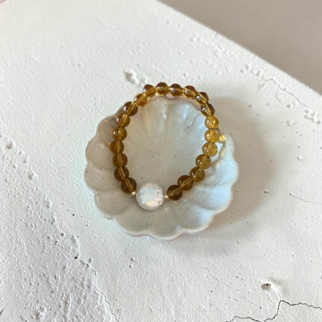 A beaded stretch bracelet with translucent amber-yellow beads and a single white pearl-like accent, displayed on a small white scalloped dish against a textured off-white surface.