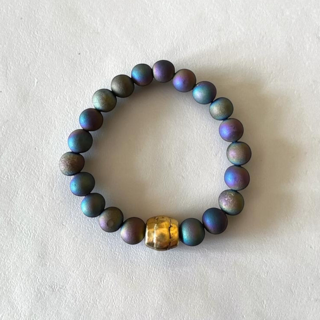 A beaded stretch bracelet made of iridescent dark blue, green, and purple pearls with a single textured gold accent bead, displayed on a textured off-white surface.