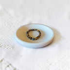 A beaded bracelet with dark brown, black, and metallic gold beads resting on a small round ceramic dish over a white textured fabric background.