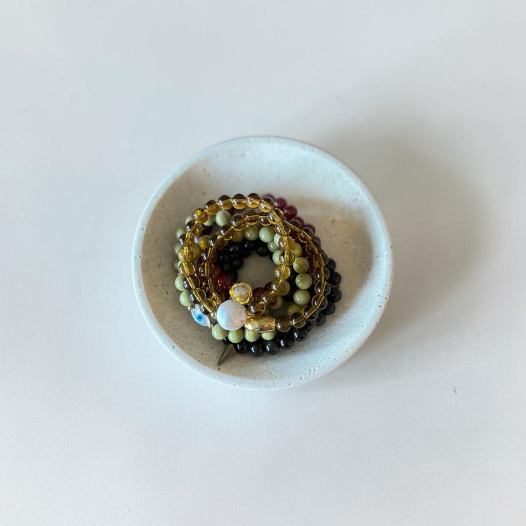 A stack of beaded bracelets with mixed colors including olive green, black, gold, and white, displayed in a small white ceramic dish on a light off-white background.