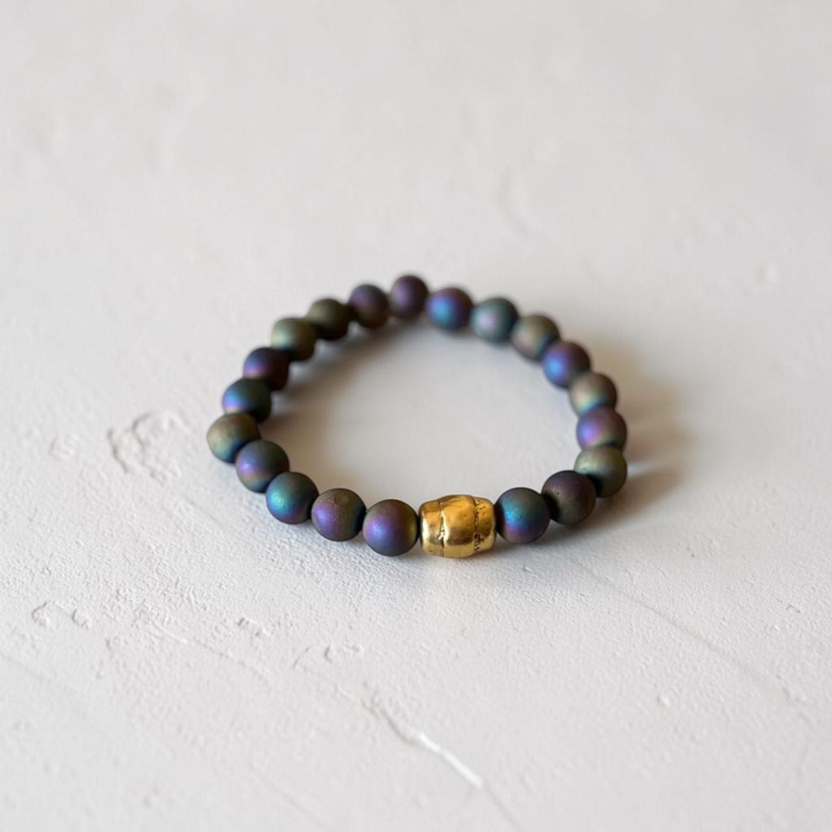 A beaded stretch bracelet featuring iridescent dark purple and blue-green beads with a single textured gold accent bead, displayed on a light textured surface.