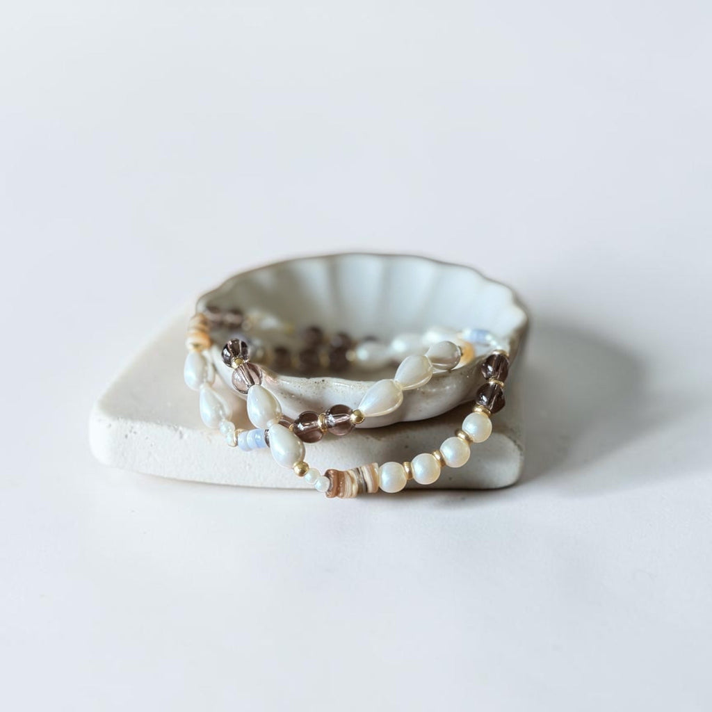 A stack of beaded bracelets with white, brown, and gold-toned beads, displayed on a small scalloped ceramic dish over a square ceramic block against a clean white background.