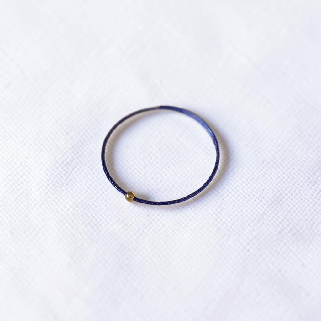 A thin purple blue wire seed bead bracelet with a small gold metal bead accent, displayed on a textured white fabric background.