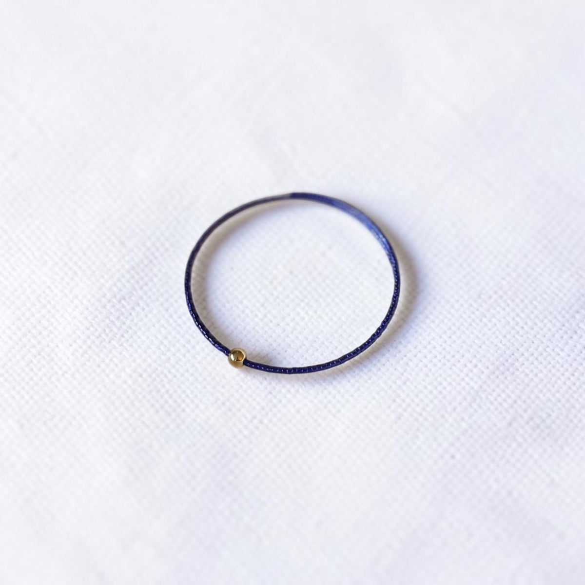 A thin purple blue wire seed bead bracelet with a small gold metal bead accent, displayed on a textured white fabric background.
