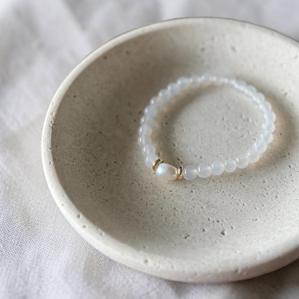 A beaded stretch bracelet with white translucent beads and a gold accent, resting inside a speckled beige ceramic dish on a light fabric surface.
