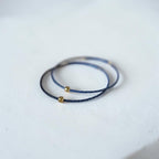 Two delicate wire bracelets, one in dark navy blue, each accented with a small gold bead, resting on a white textured surface.