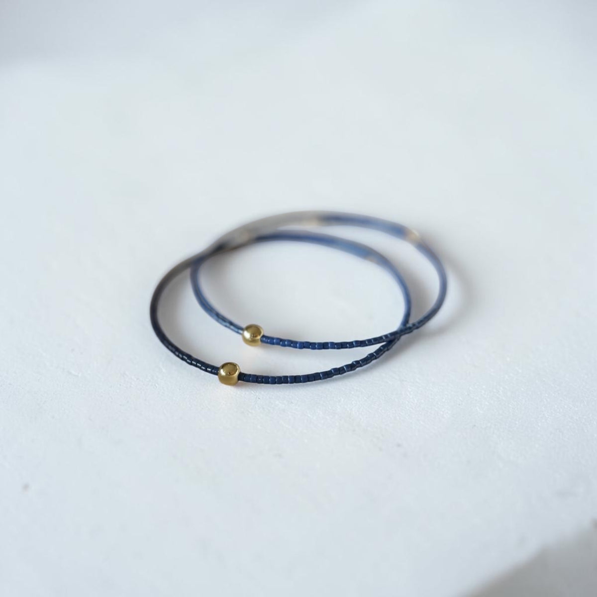 Two delicate wire bracelets, one in dark navy blue, each accented with a small gold bead, resting on a white textured surface.